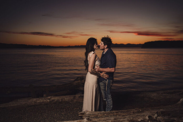 Sunset beach engagement photos of a couple standing on driftwood along Puget Sound in Washington