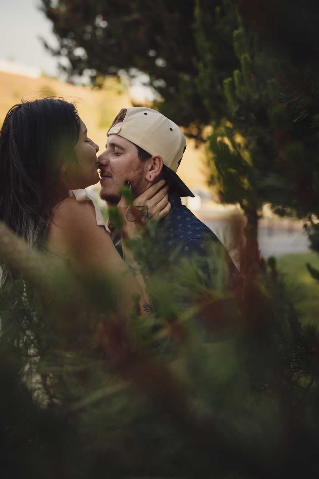 Engaged couple sharing a kiss beneath the pines at Chambers Bay in University Place