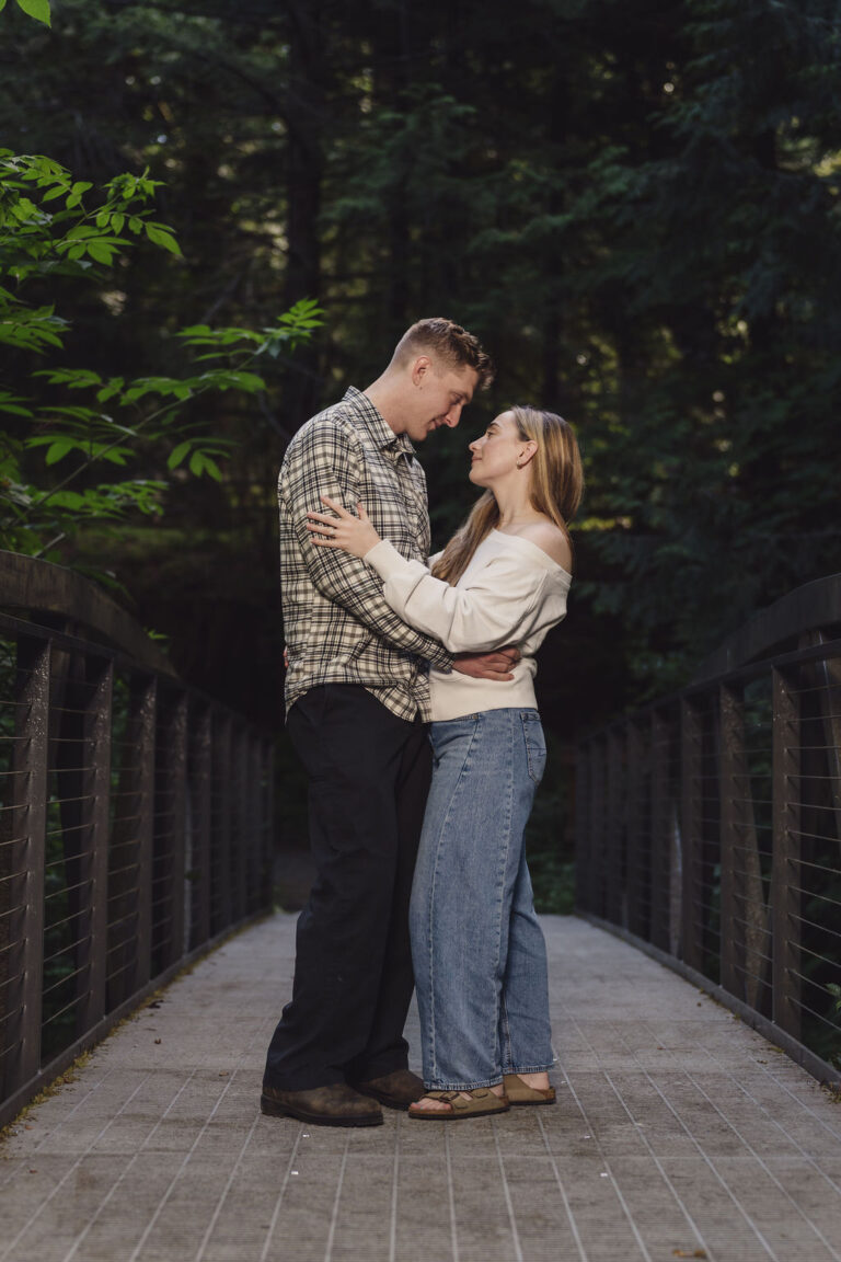 Engaged couple embracing on the footbridge beneath the evergreens at Scenic Beach State Park