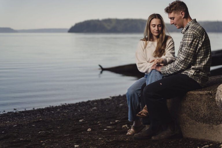 Engaged couple cuddling on driftwood by the water during Seabeck engagement photos