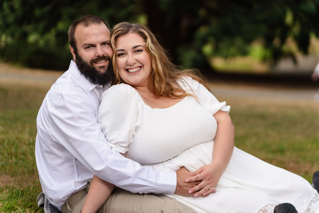 Engaged couple snuggled together on the lawn smiling at the camera at Point Defiance in Tacoma