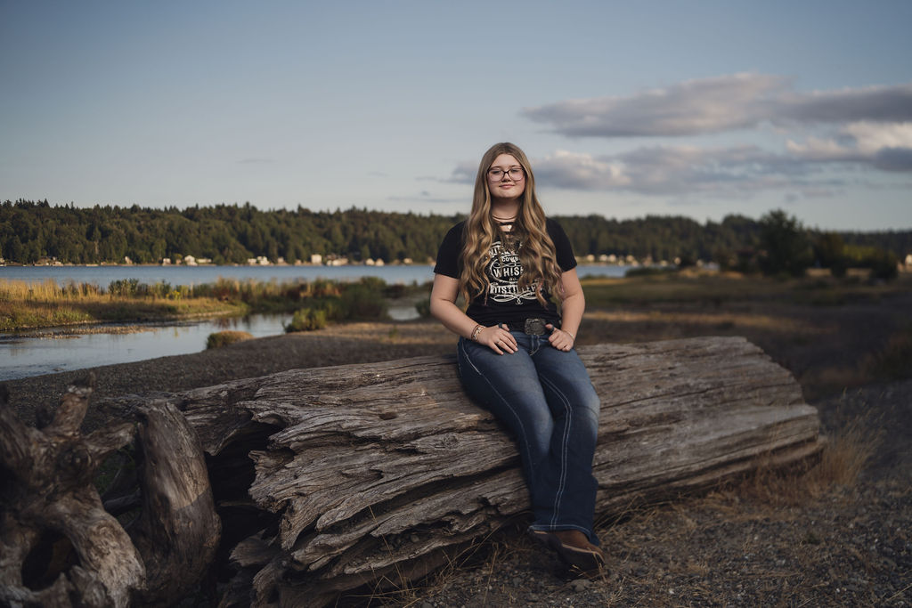 Senior sitting on a massive weathered driftwood log with the Hood Canal shoreline stretching behind her