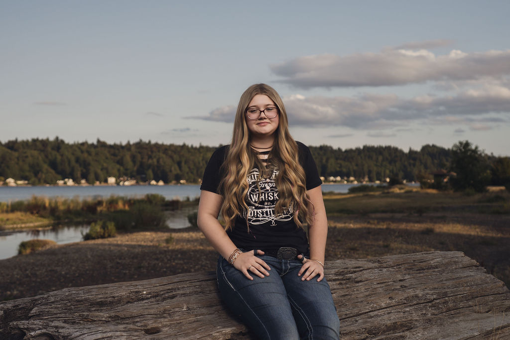 Senior sitting on a driftwood log with the wide Hood Canal estuary and far shoreline behind her at Belfair State Park