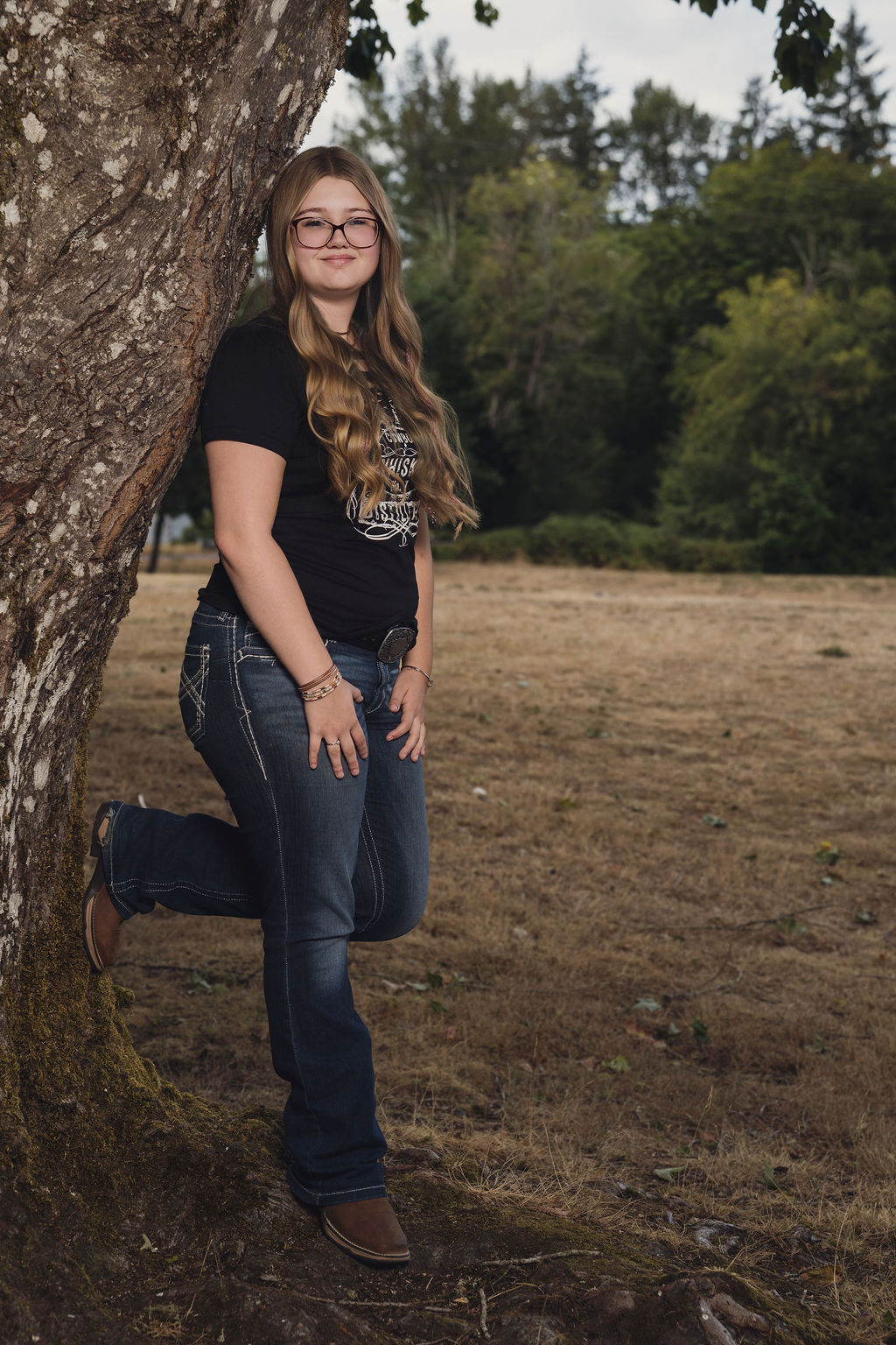 Senior in a western graphic tee and jeans with one boot propped against a big maple at Belfair State Park