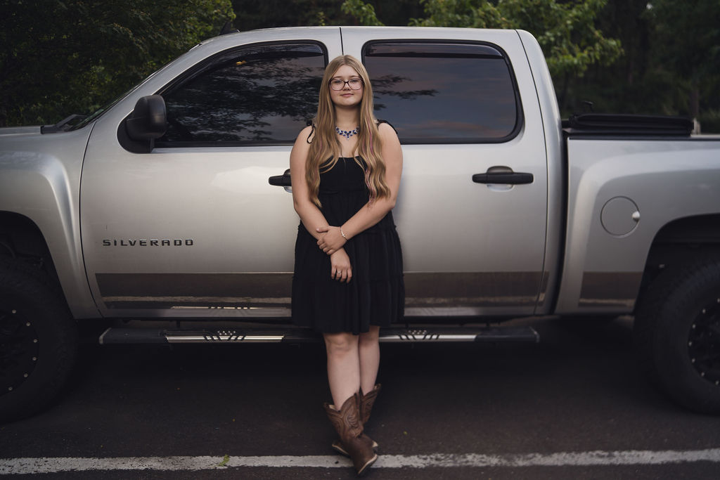 Senior in a black dress and cowboy boots standing beside her silver Chevy Silverado in Belfair