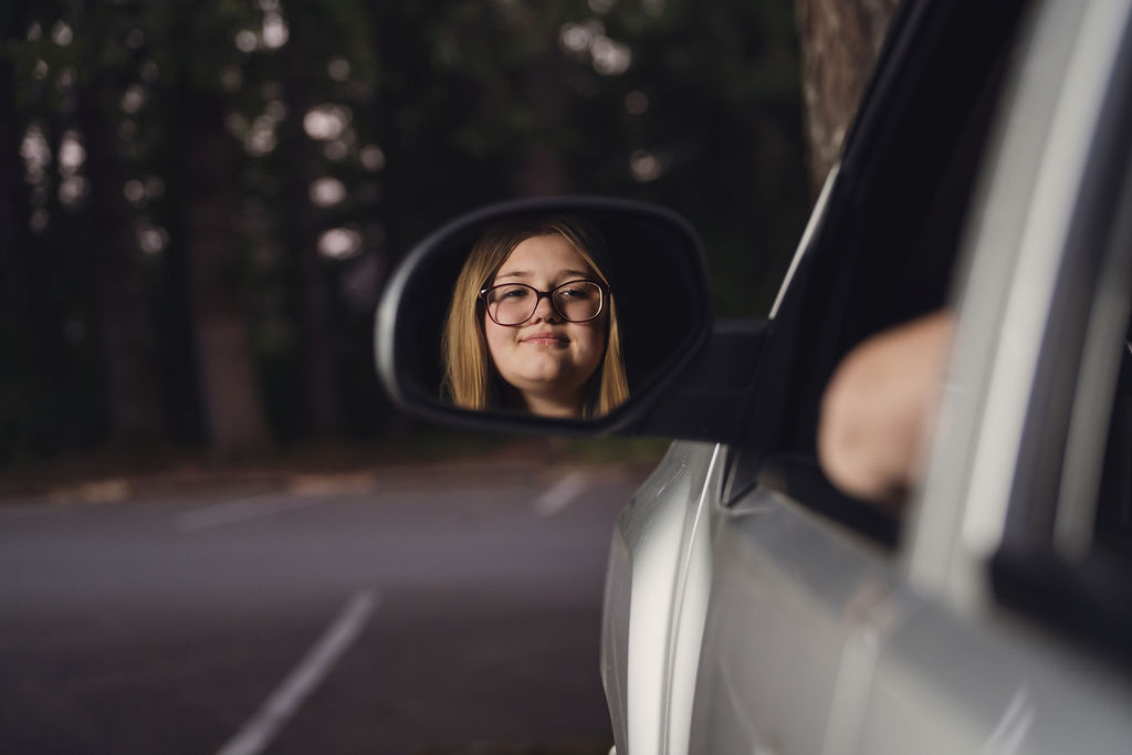 Senior girl's face framed in the side mirror of her silver pickup on a tree lined road in Belfair