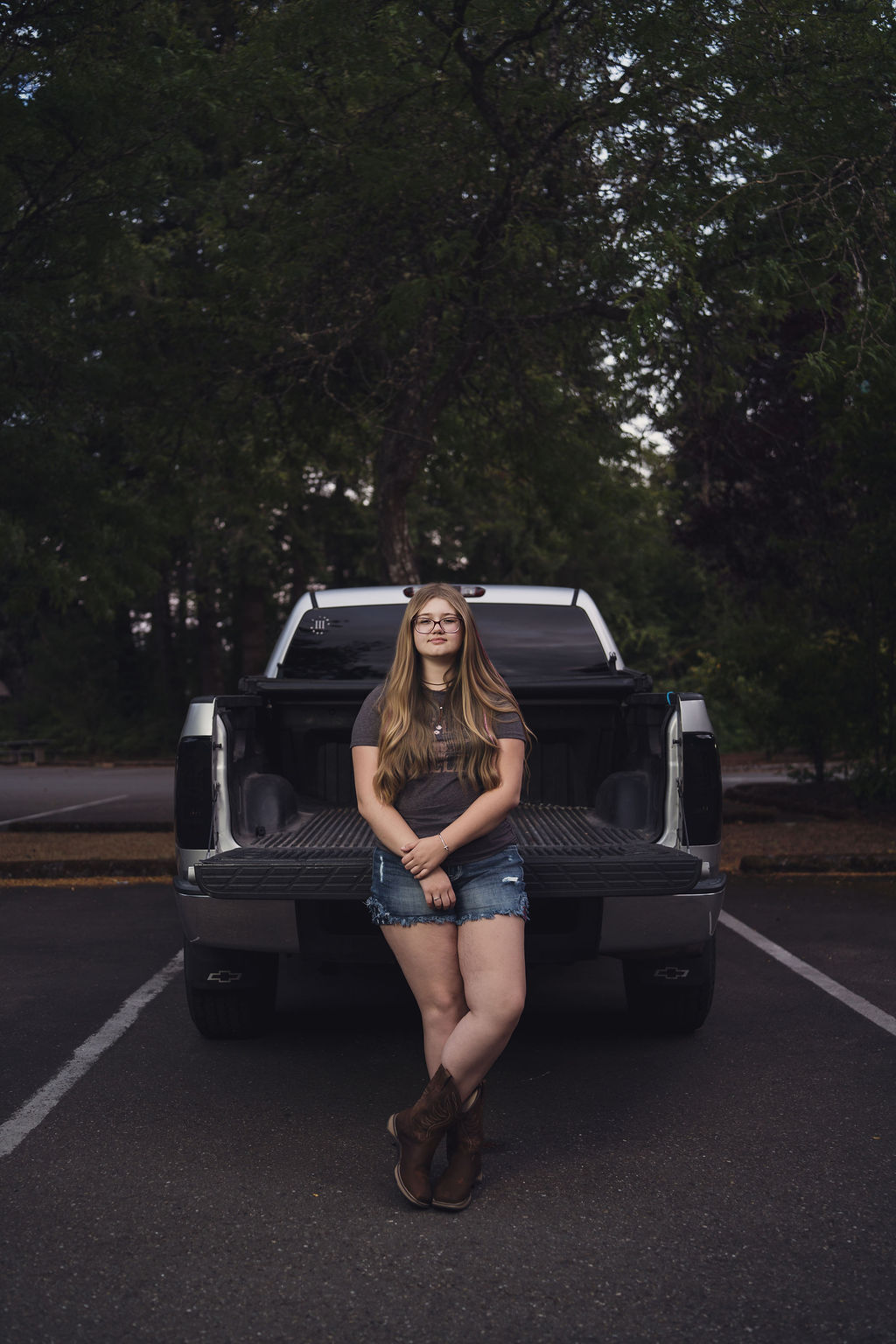 Senior seated square on the tailgate of her silver pickup framed by evergreens near Hood Canal