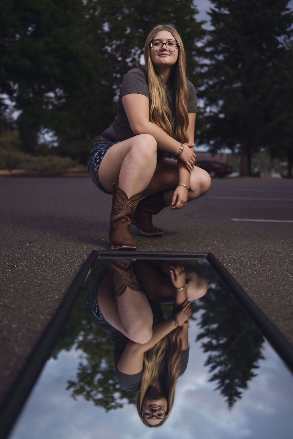 Creative senior photo crouched over a mirror on the pavement reflecting her portrait and the evening sky in Belfair
