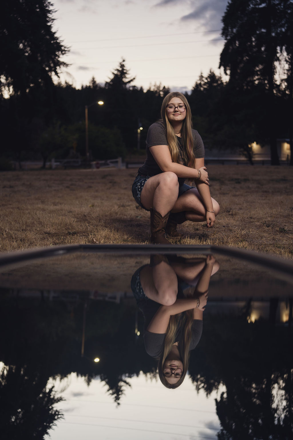 Senior crouched at dusk with a ground mirror reflecting her silhouette and the evening sky in Mason County