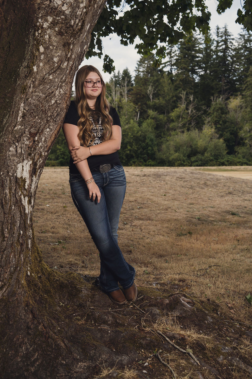 Full length photo of a senior in jeans and boots standing at the base of a maple with a dry summer field behind her