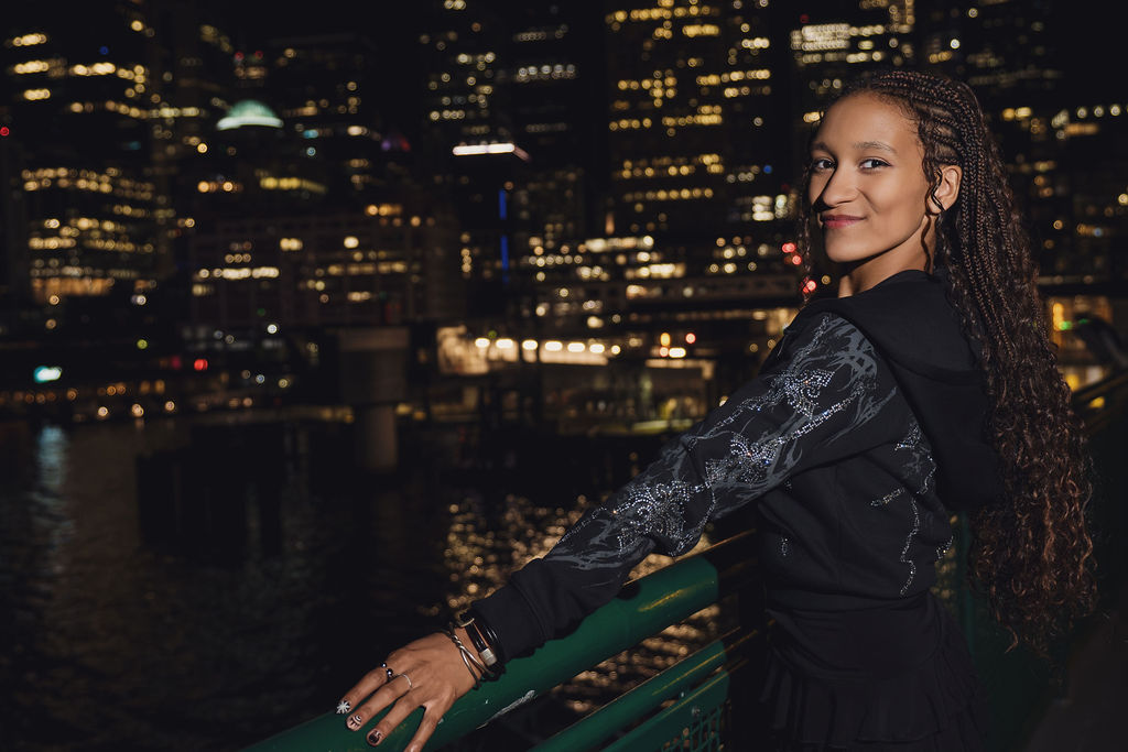 High school senior glancing back with a smile while holding the green ferry railing as downtown Seattle waterfront piers glitter across the dark water behind her.