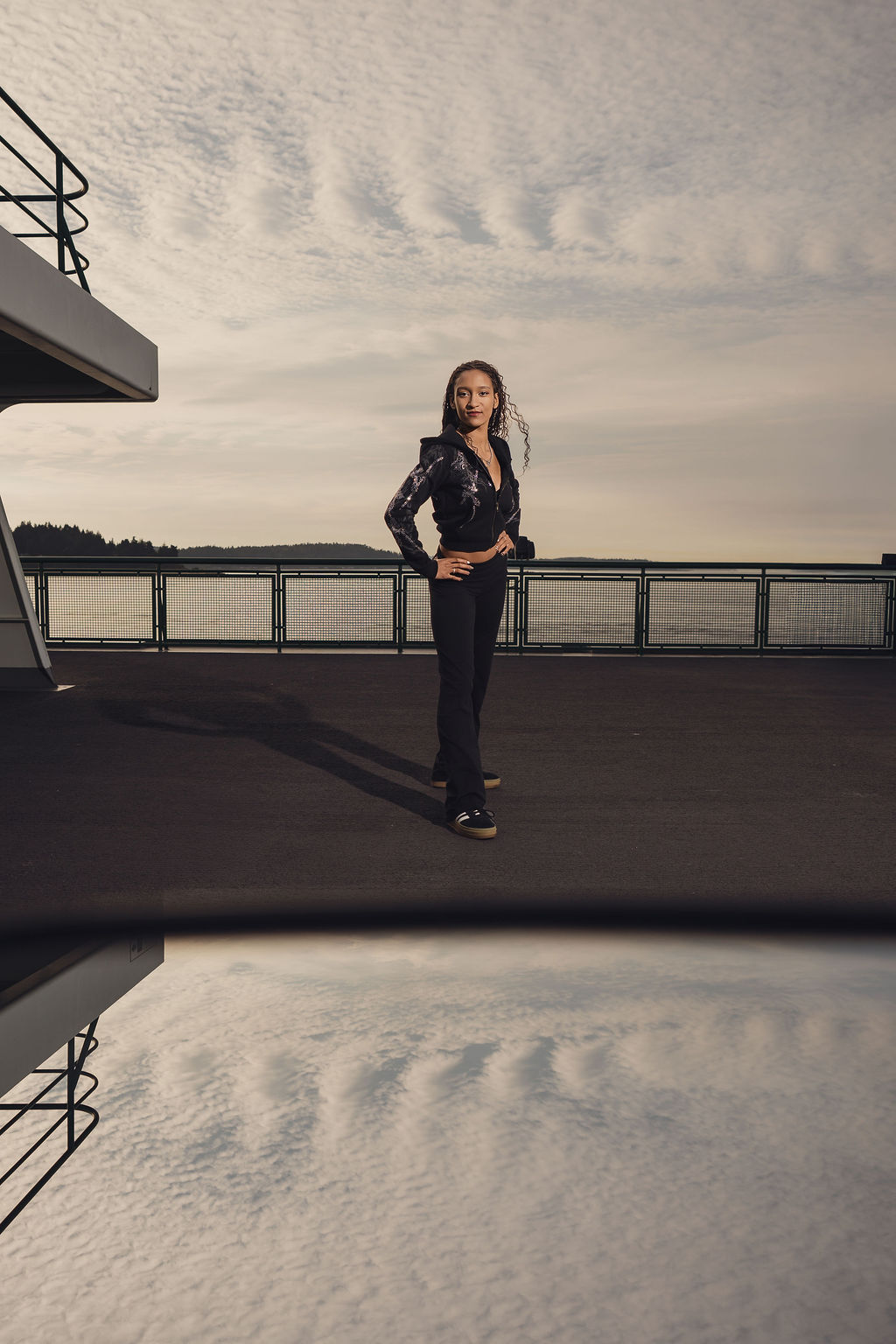 Full length photo of a high school senior standing hands on hips on the top ferry deck under a dramatic cloud filled sky mirrored in a reflection, Bremerton senior pictures