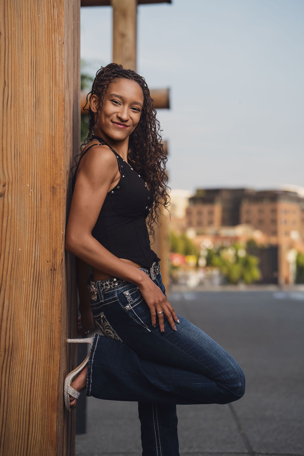 High school senior in a studded black top and jeans leaning against a large wooden pier post with brick buildings blurred behind her for Seattle waterfront senior pictures