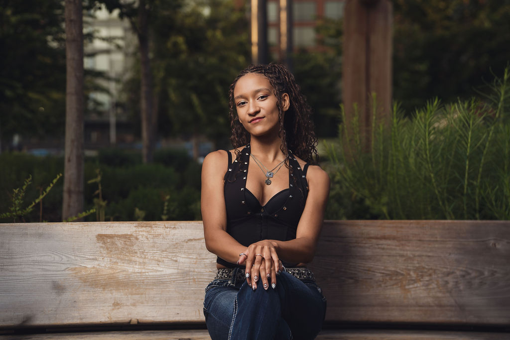 High school senior seated on a wooden park bench with hands resting on her knee and lush greenery behind her, senior portraits near the Seattle waterfront