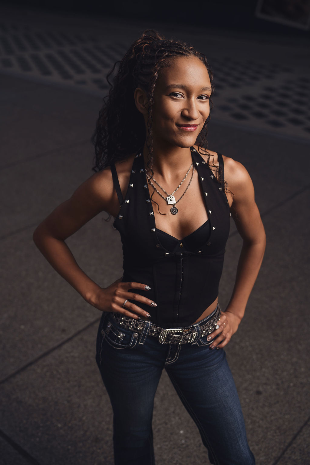 High school senior smiling with hands on her hips in a corset top and jeans on a downtown Seattle plaza lit by patterned lights during her senior session
