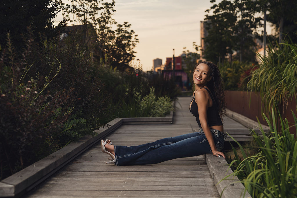 High school senior sitting on a wooden boardwalk edge between tall grasses with legs extended as the sun sets over the Seattle waterfront park