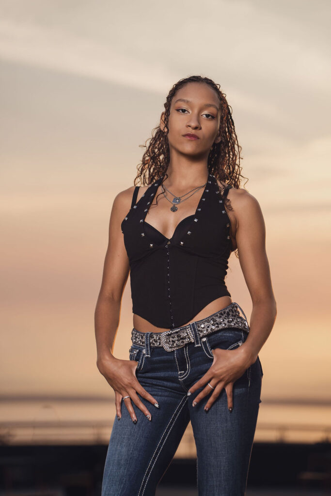 High school senior in a studded black corset top and jeans posing with hands at her hips against a peach sunset sky during a Seattle waterfront senior session