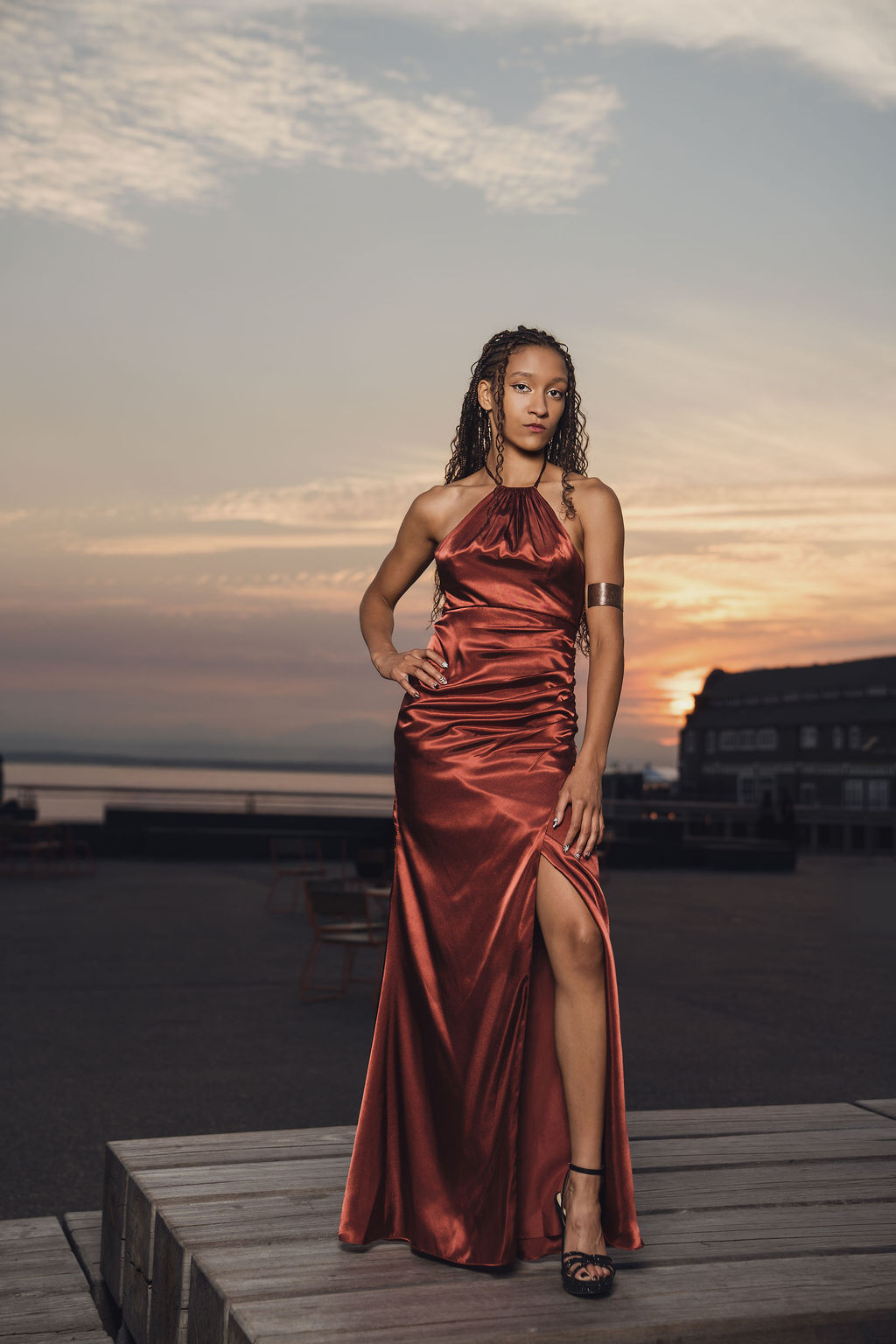 High school senior in a copper satin halter gown with a gold arm cuff standing on a wooden platform at sunset during Seattle waterfront senior pictures