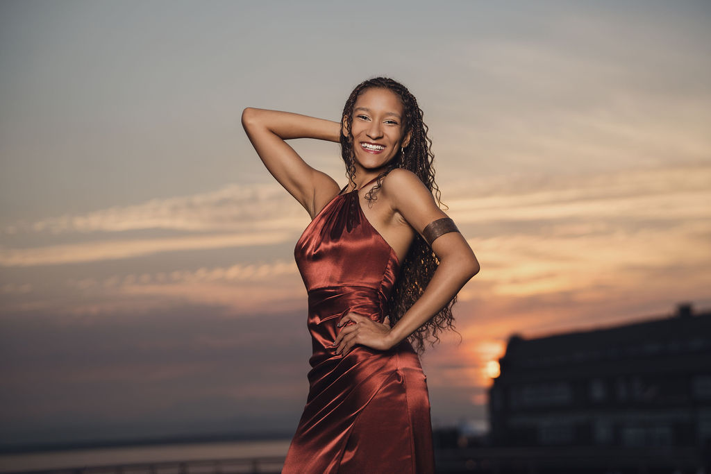 Senior laughing with one hand behind her head in a copper satin gown, photographed against a glowing sunset sky during a Puget Sound senior session