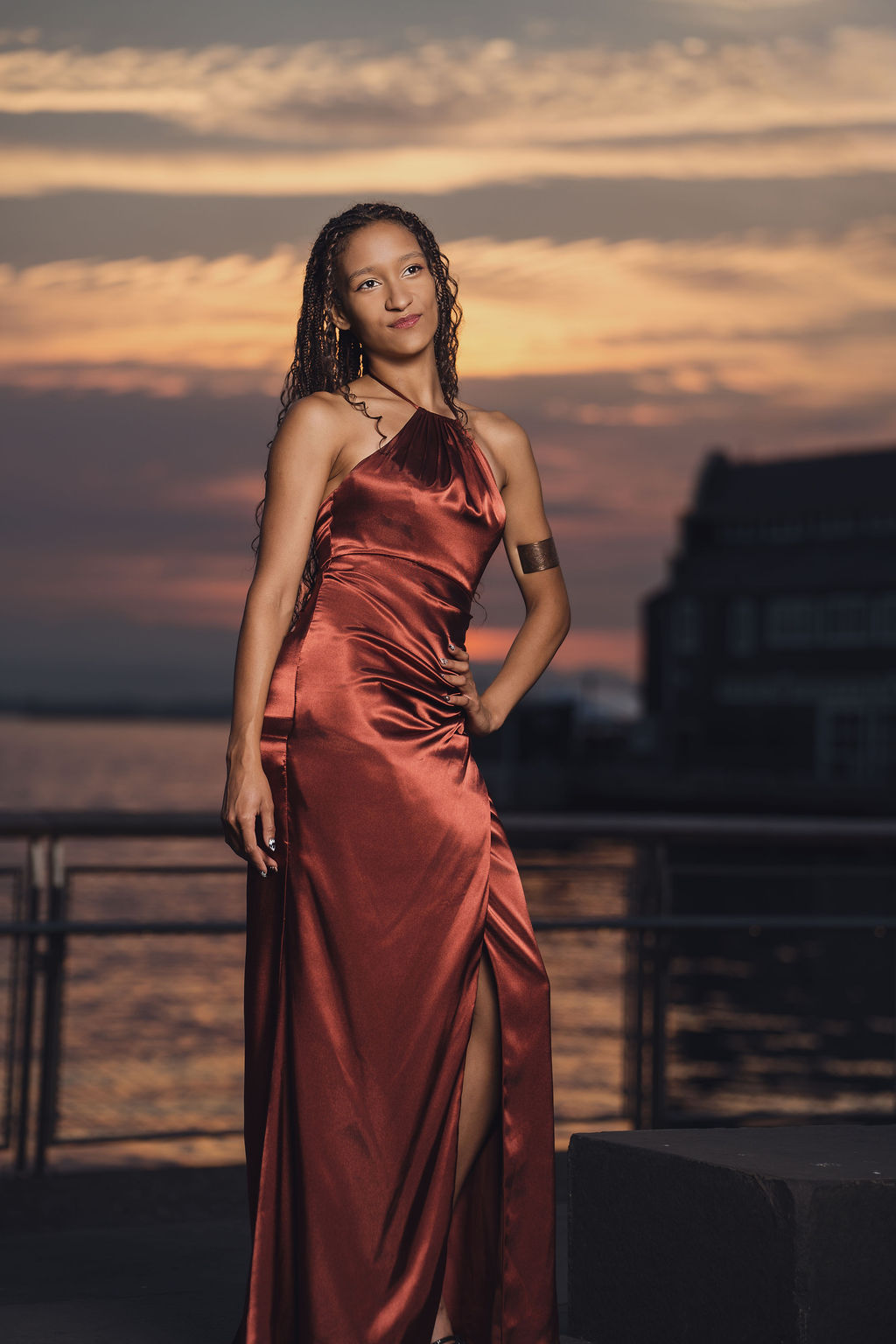 Senior in a satin halter gown smiling softly beside a waterfront railing as sunset colors reflect on the water during a Seattle senior photographer session