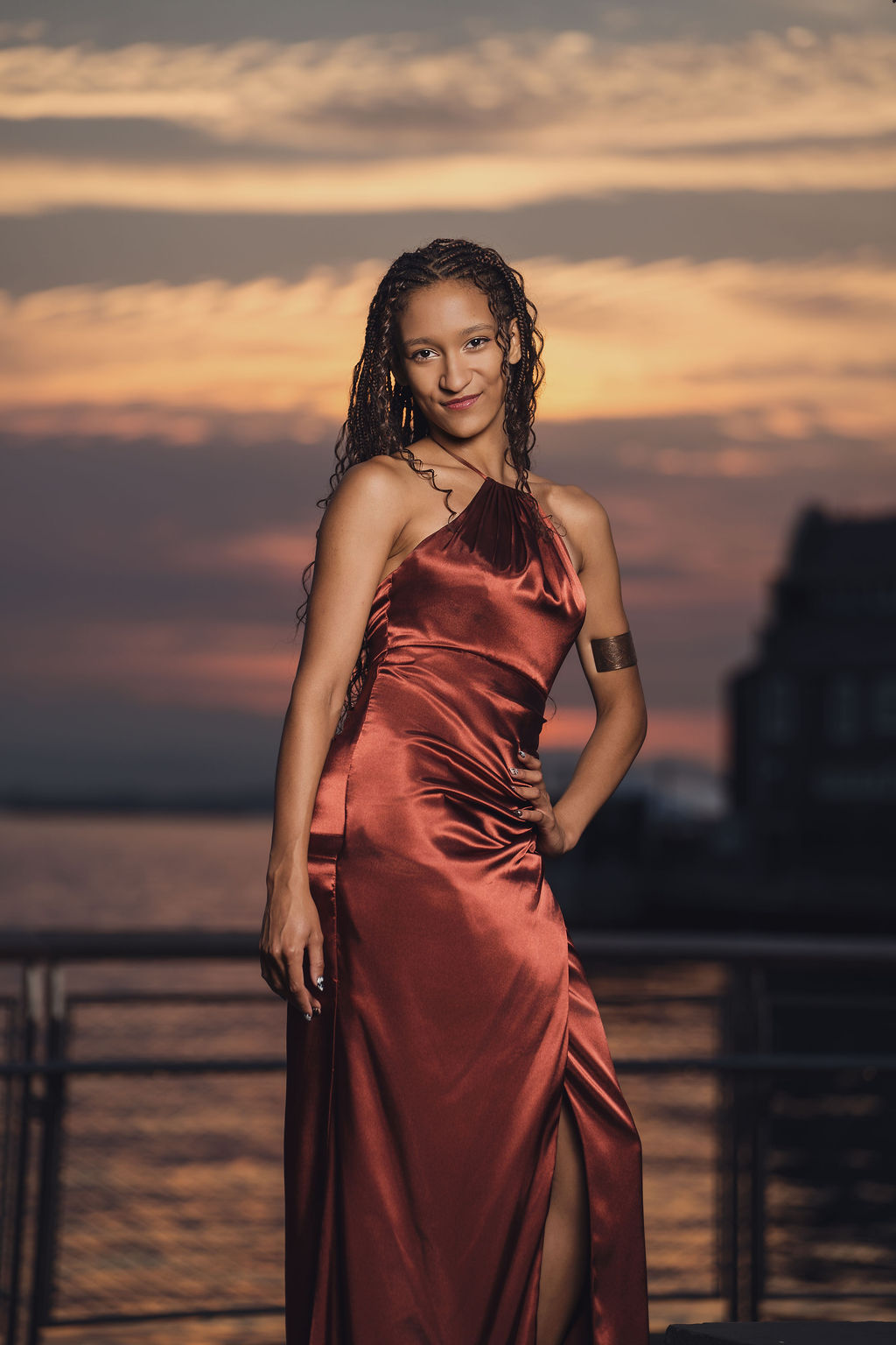 Senior wearing a copper satin dress and cuff bracelet smiling at the camera with the sunset glowing over Puget Sound water behind her