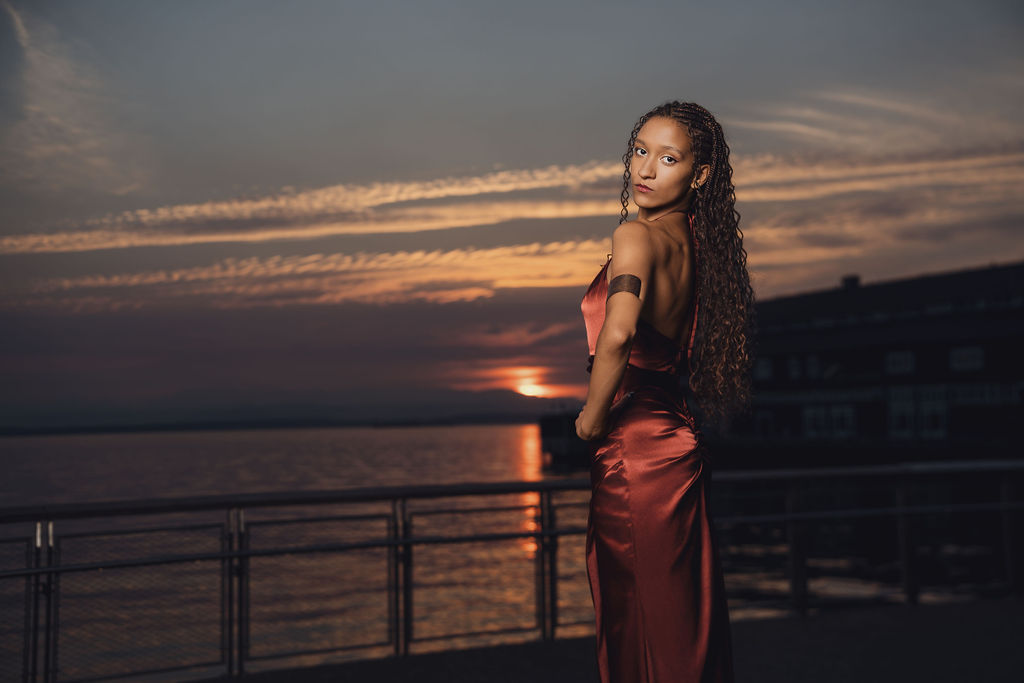 Moody portrait of a high school senior glancing back over her bare shoulder as the sun touches the horizon across Puget Sound from the Seattle waterfront