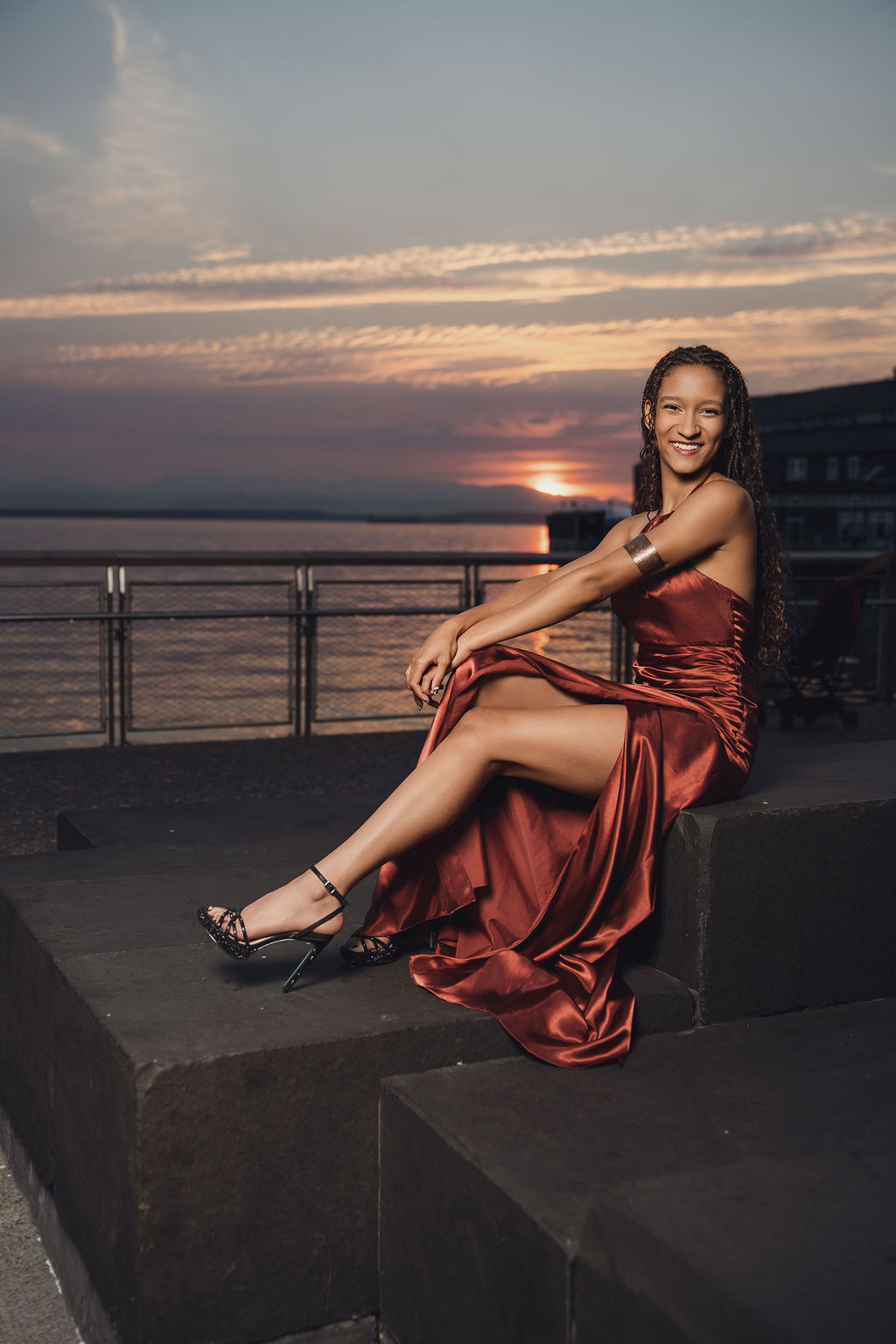 Smiling senior in a copper satin gown seated on stone steps with legs crossed, her dress draped over the edge as the sun sets over the water in Seattle