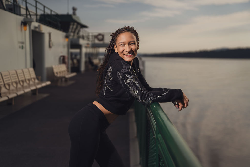 Senior laughing with a wide smile while leaning on the green railing of a Washington State Ferry, with still water and distant hills behind her evening session.