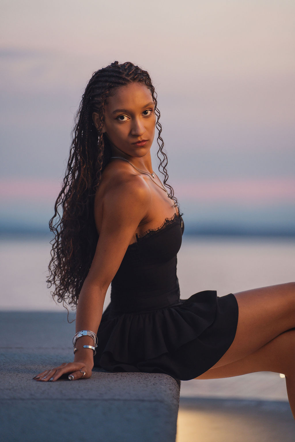 High school senior in a black dress seated on a concrete ledge looking over her shoulder with a pastel sunset over Puget Sound behind her.