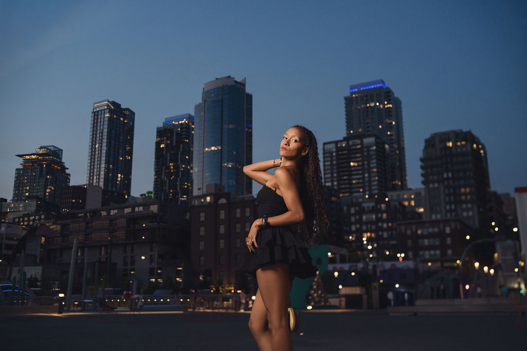 Full length photo of a high school senior posing in a plaza with the lit Seattle skyline stretching across the twilight sky, Seattle waterfront senior pictures.