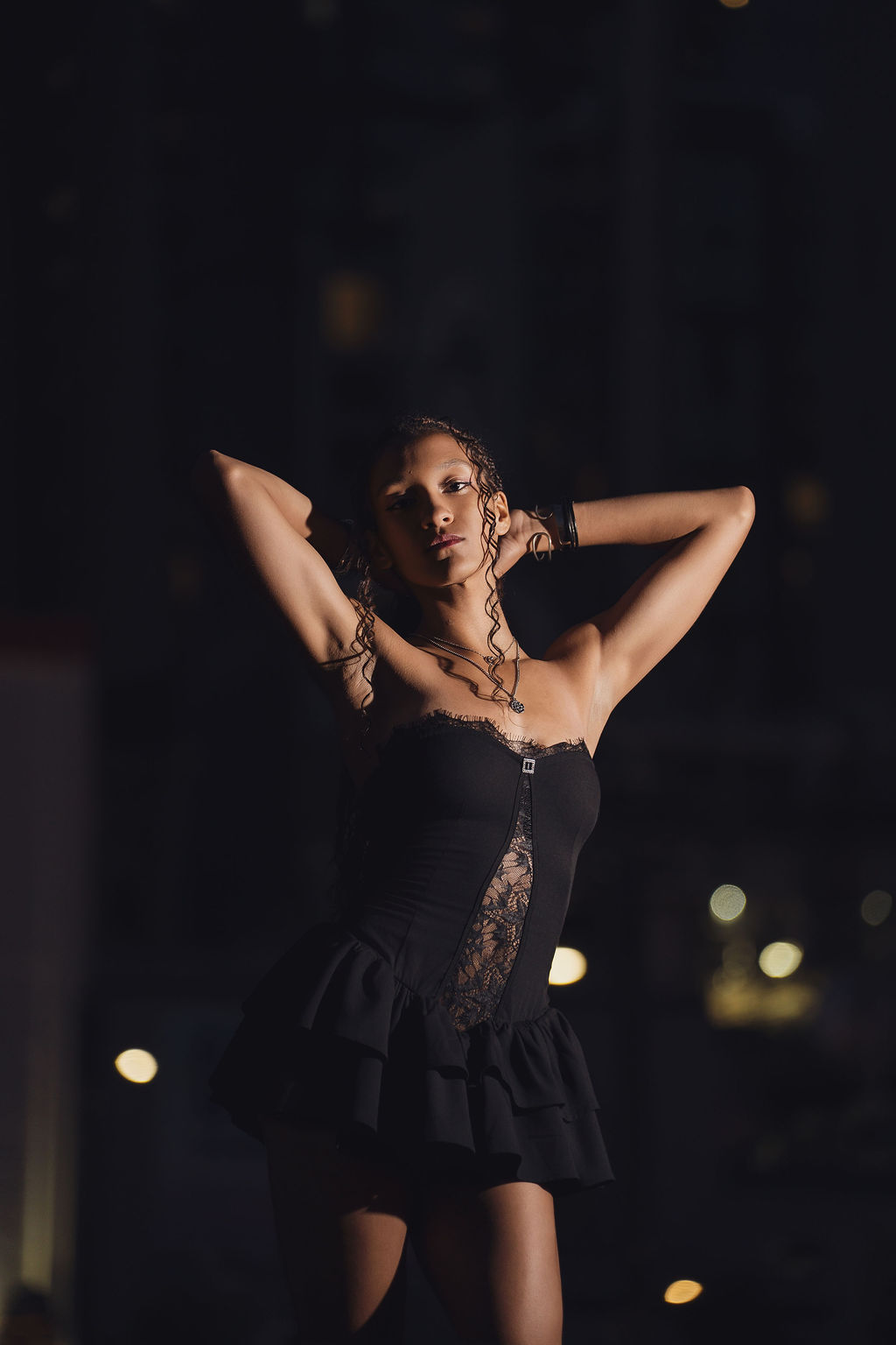 Moody close portrait of a high school senior with arms lifted behind her head, a single beam of light sculpting her against the dark city night.