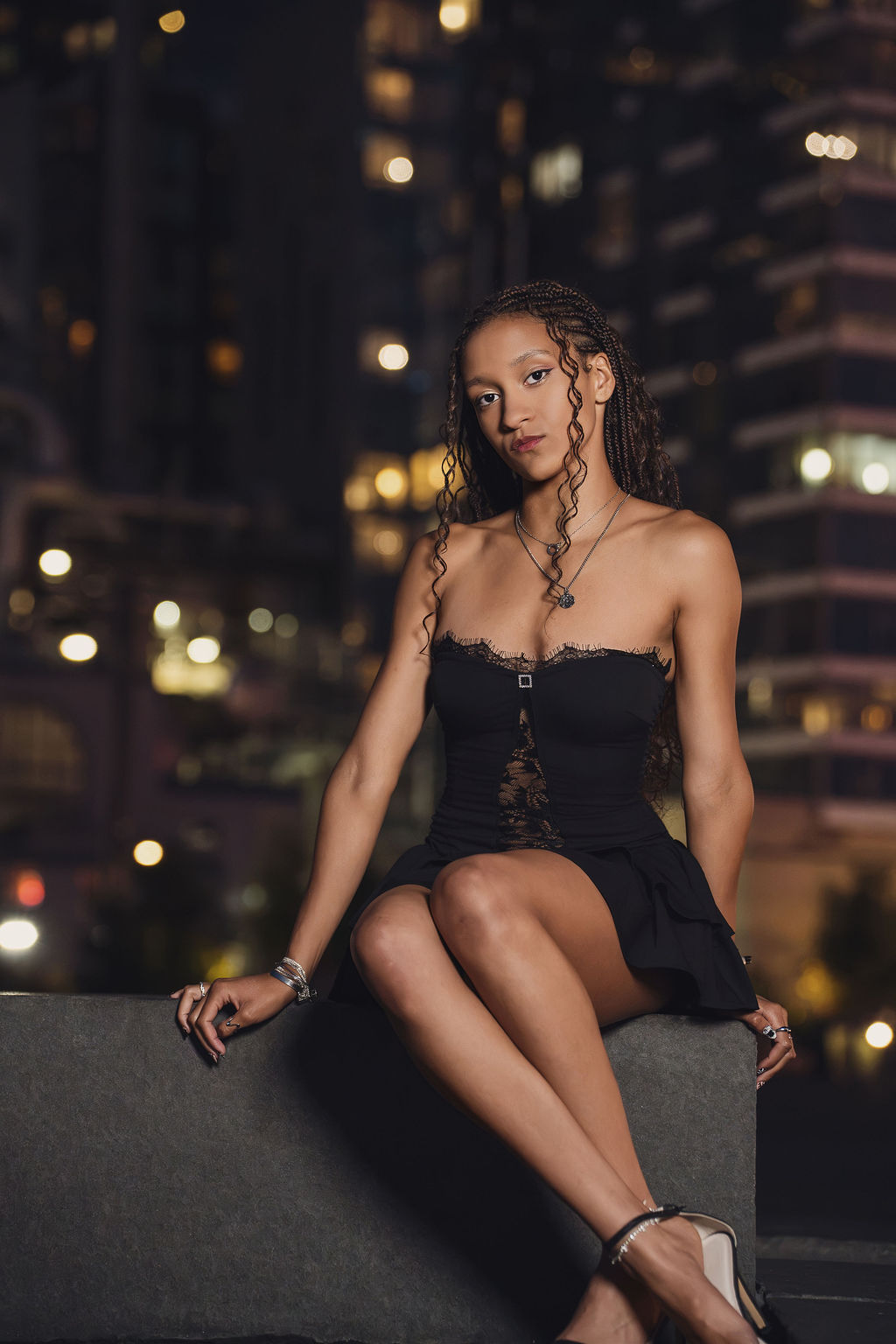 High school senior in a black lace trimmed dress sits on a concrete ledge with glowing Seattle high rise windows behind her during a night portrait session