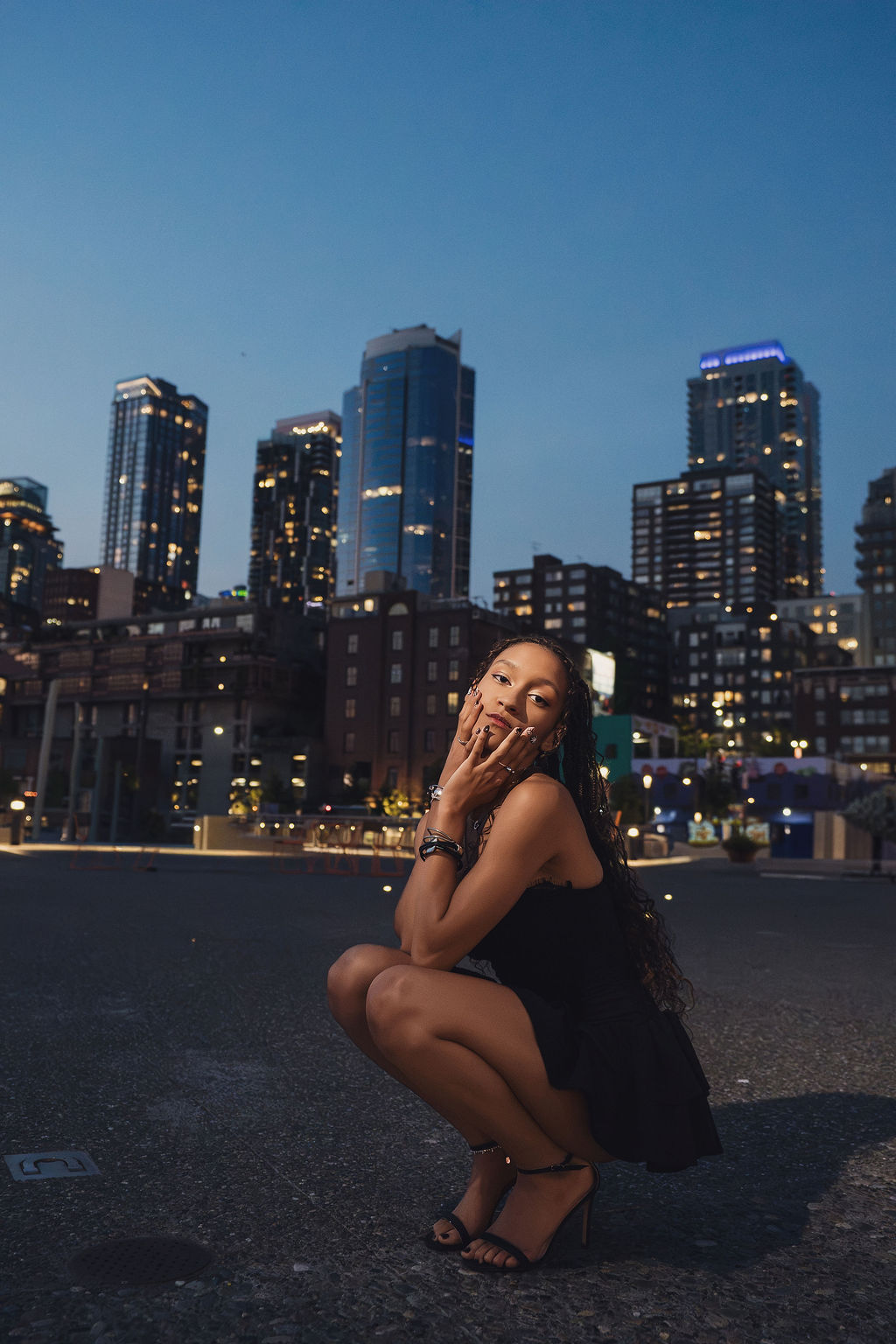 High school senior crouches on a downtown street with hands framing her face as the lit Seattle skyline rises against a deep blue dusk sky during her senior session