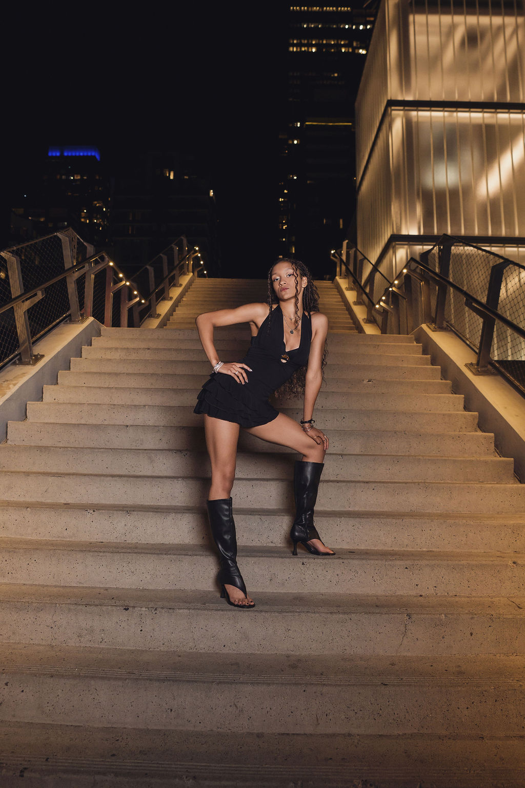 Senior in tall black boots poses with one leg extended down a wide concrete staircase lit by string lights and a glowing glass building in downtown Seattle at night