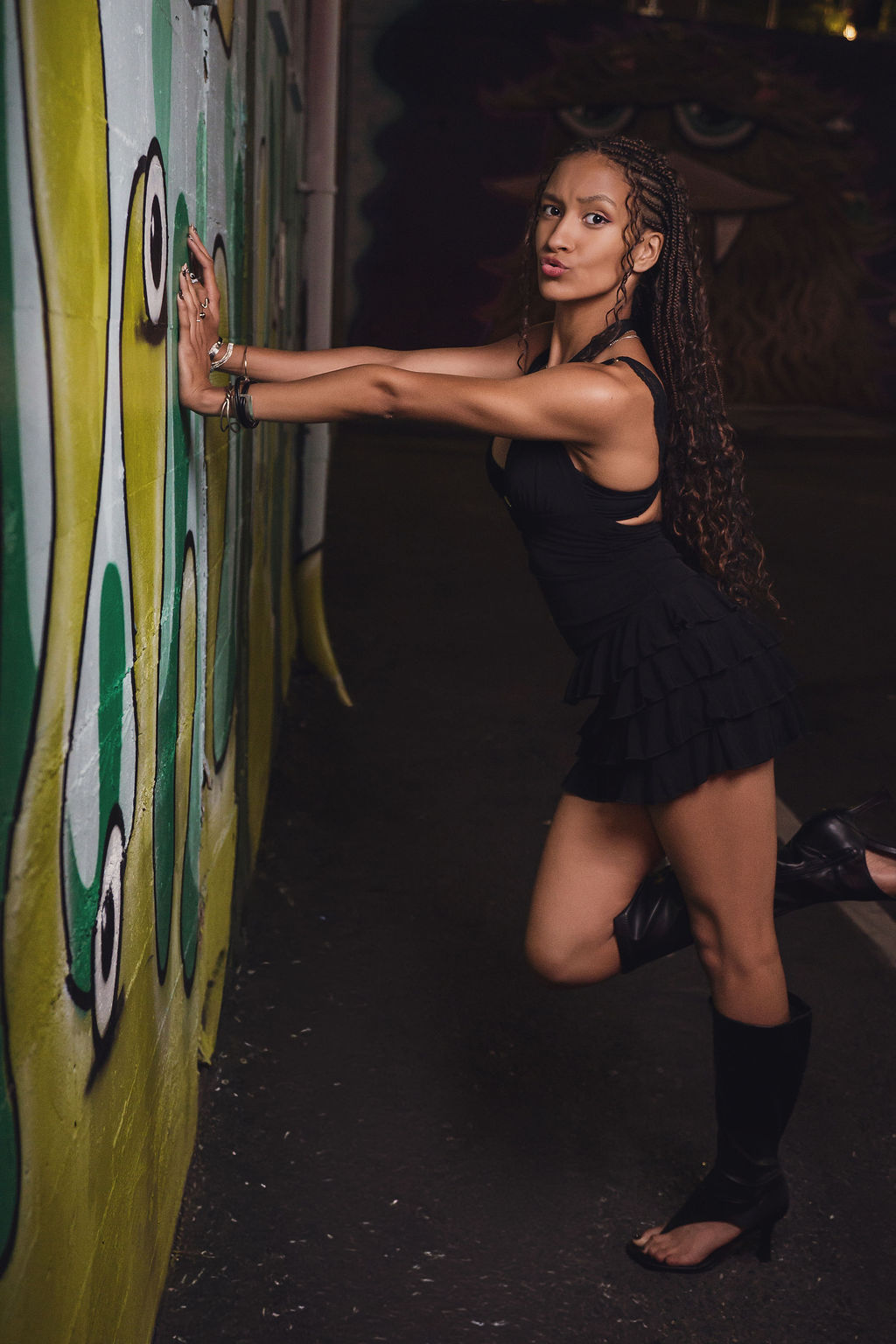 High school senior makes a playful kissy face while pressing her hands against a green graffiti mural and kicking one boot up, a fun stop on her Seattle senior session