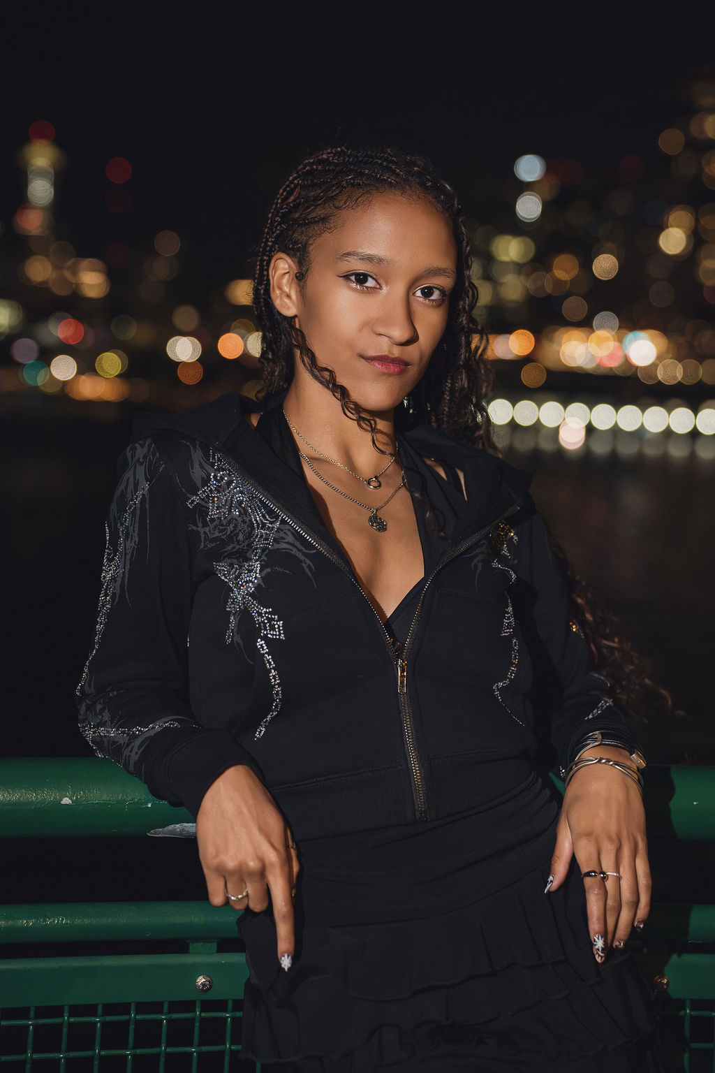 High school senior in a black rhinestone hoodie leaning back on a green ferry railing with Seattle city lights glowing in soft bokeh behind her during nighttime Seattle waterfront senior pictures.