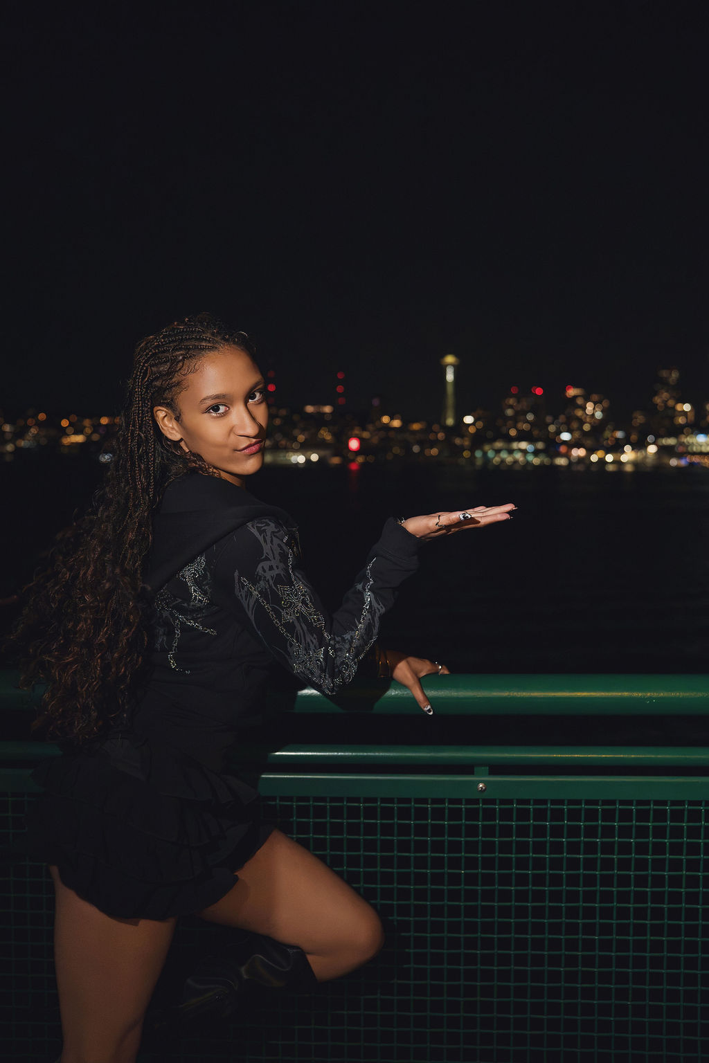Senior holding the green ferry railing and extending an open hand toward the Space Needle glowing on the night skyline, a playful moment from a Puget Sound senior session.