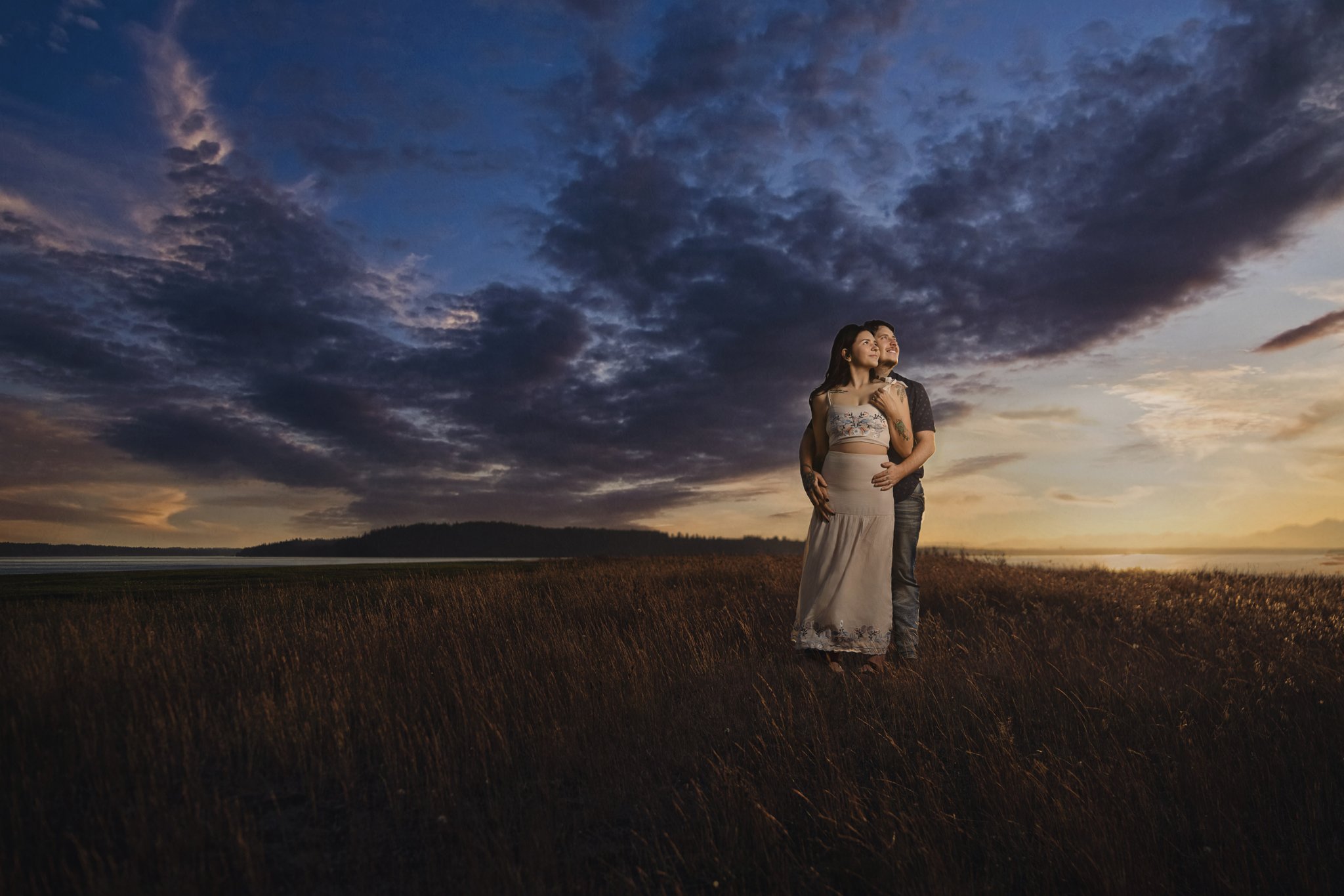 Signature edit of an engaged couple in the golden grass at dusk at Chambers Bay with storm clouds parting over Puget Sound