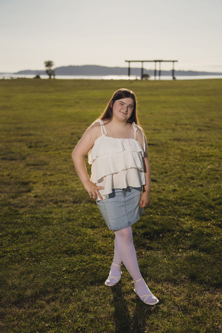 Tacoma senior photographer portrait of a senior in a white ruffled top and denim skirt standing in the grass above Puget Sound at Chambers Bay