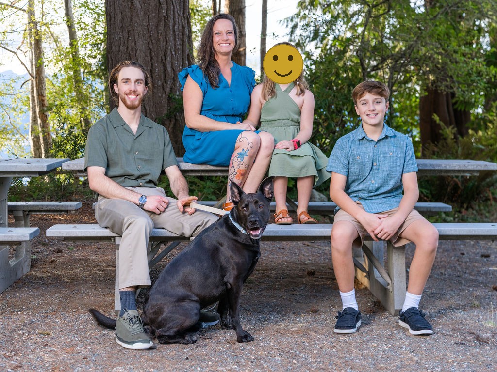 Family of four with their dog seated at a picnic table in the woods, faces covered for privacy