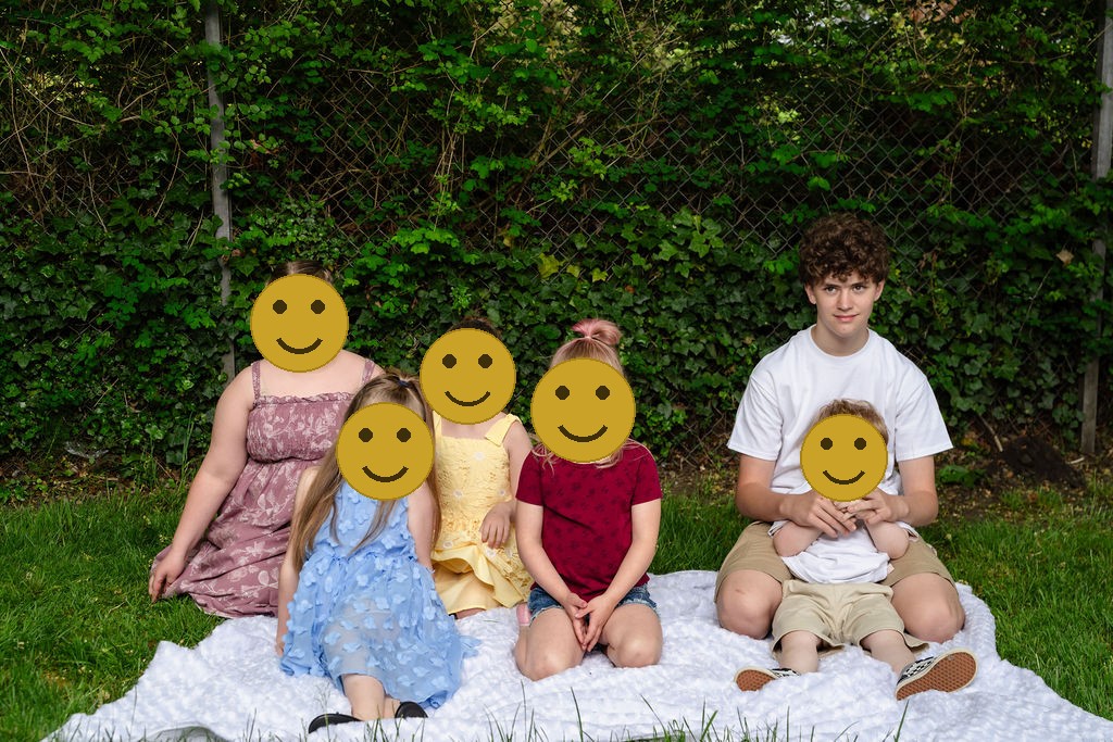 Six cousins sitting together on a white blanket in front of leafy greenery at a Kitsap County park, faces covered for privacy