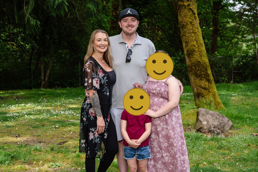 Family of four with two daughters posing together by a mossy tree at a green Kitsap County park, faces covered for privacy