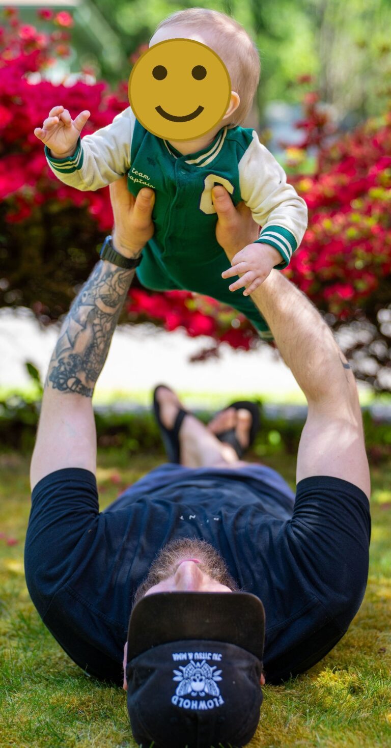 Father lying in the grass lifting his smiling baby overhead with red spring blooms behind them in Kitsap County