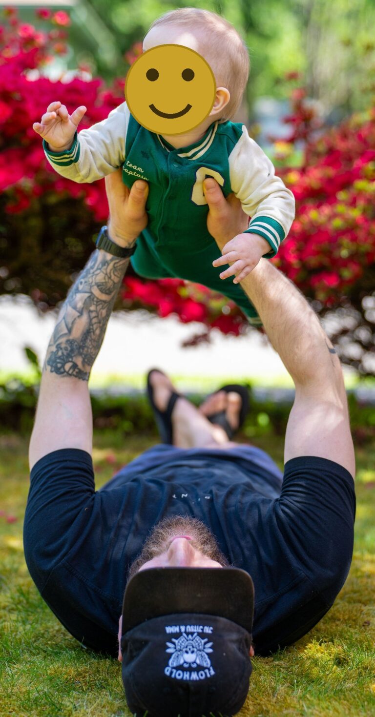 Father lying in the grass lifting his smiling baby overhead with red spring blooms behind them in Kitsap County