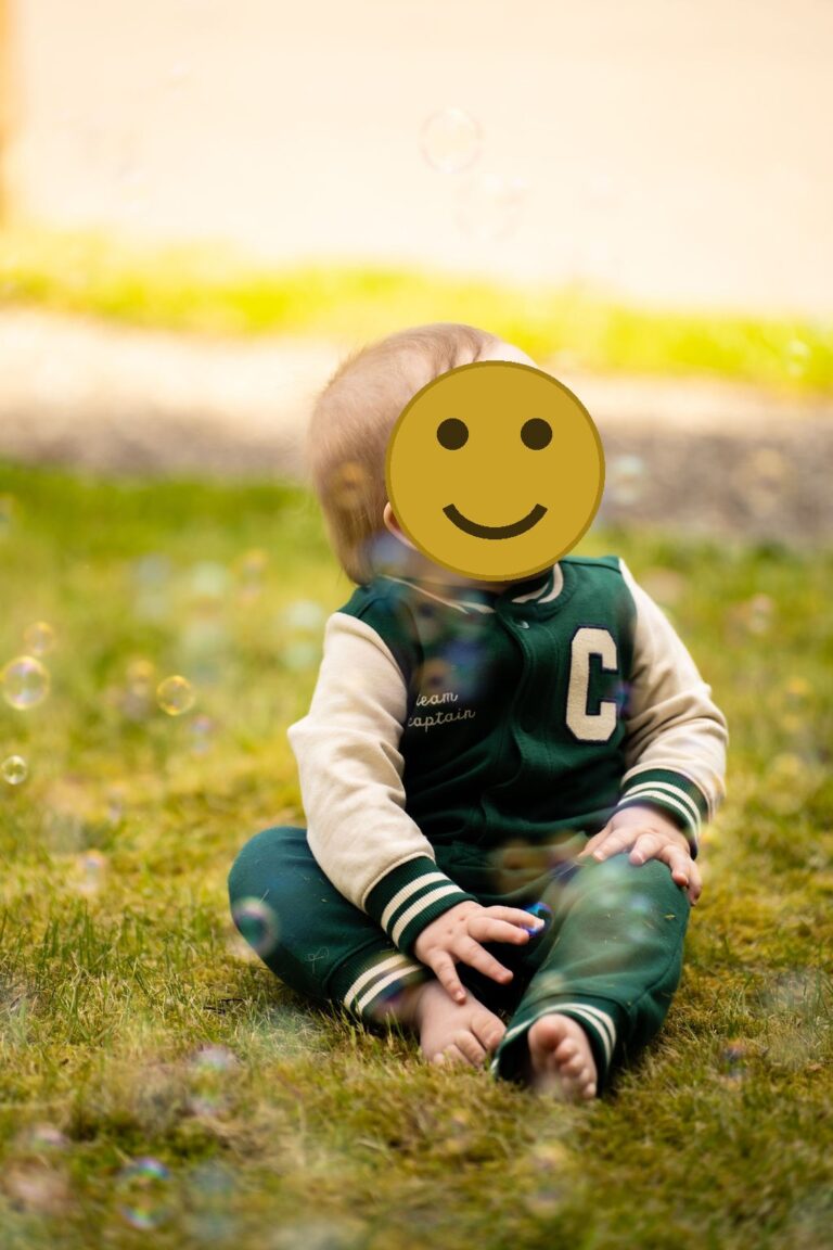 Smiling baby in a green varsity romper sitting in the grass with bubbles floating around in Kitsap County