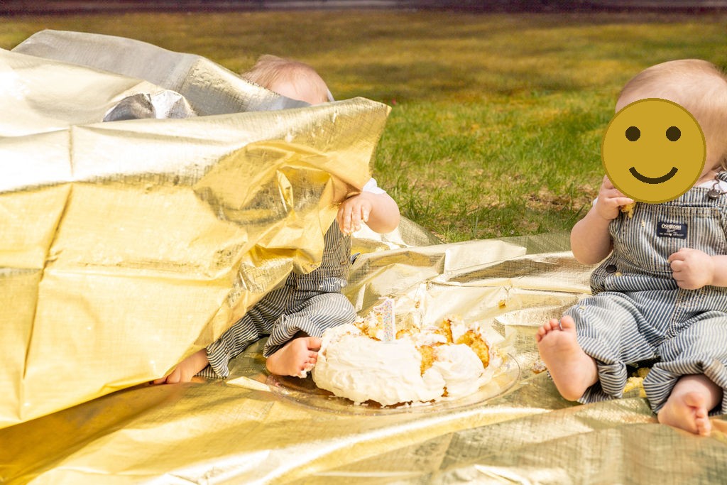 Twin babies at an outdoor first birthday cake smash, one peeking from under a gold blanket, faces covered for privacy
