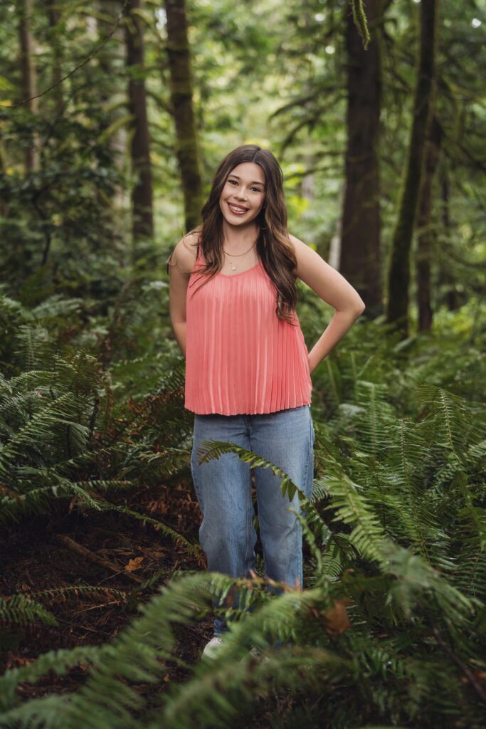 High school senior in a coral pleated top standing among sword ferns in a mossy Kitsap County forest