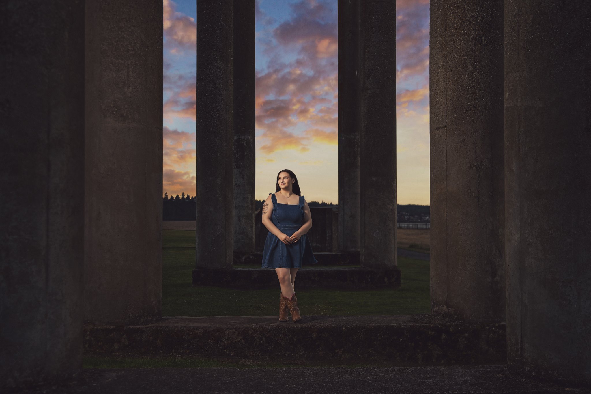 High school senior in a denim dress and cowboy boots standing among the concrete ruins columns at Chambers Bay at sunset Captured by Tacoma senior photographer Nautical Moments Photography.