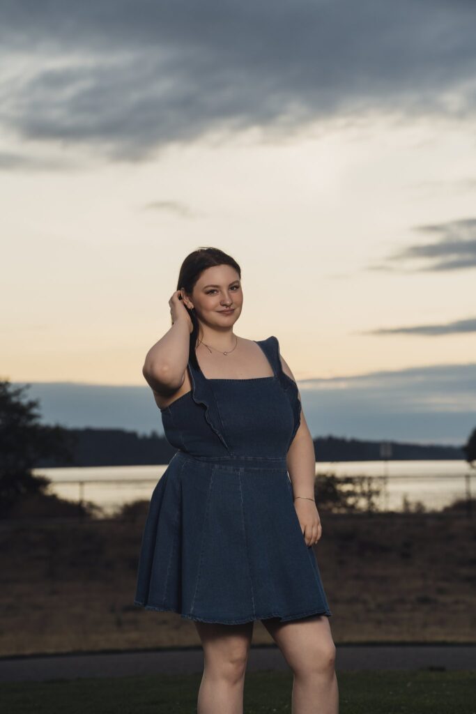 High school senior in a denim dress with Puget Sound behind her at dusk during a Chambers Bay senior session