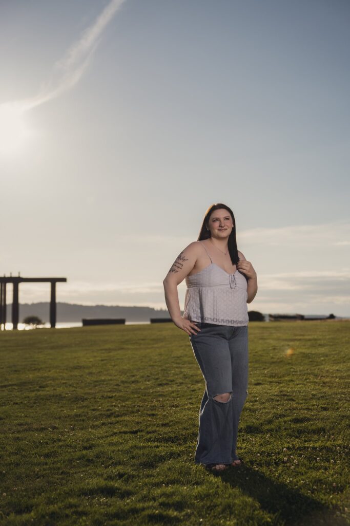 High school senior in a white lace top and jeans on the great lawn at Chambers Creek Regional Park at golden hour